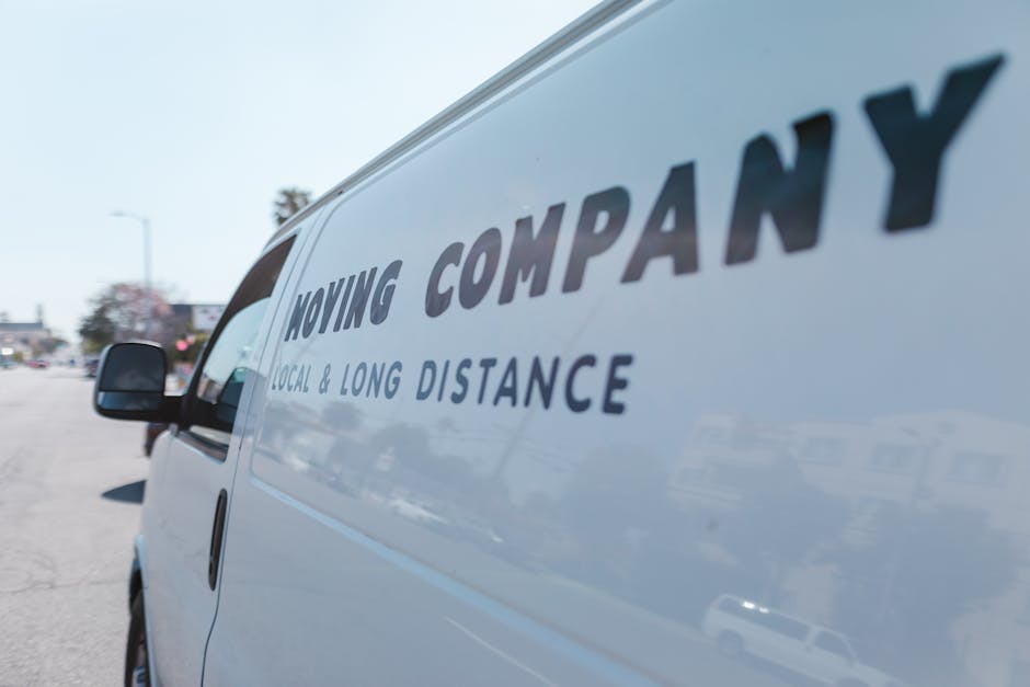 Close-up view of the side of a white moving van parked outdoors, displaying the company logo and text 'Moving Company' and 'Local & Long Distance' in bold, black letters. The van is positioned on a pa