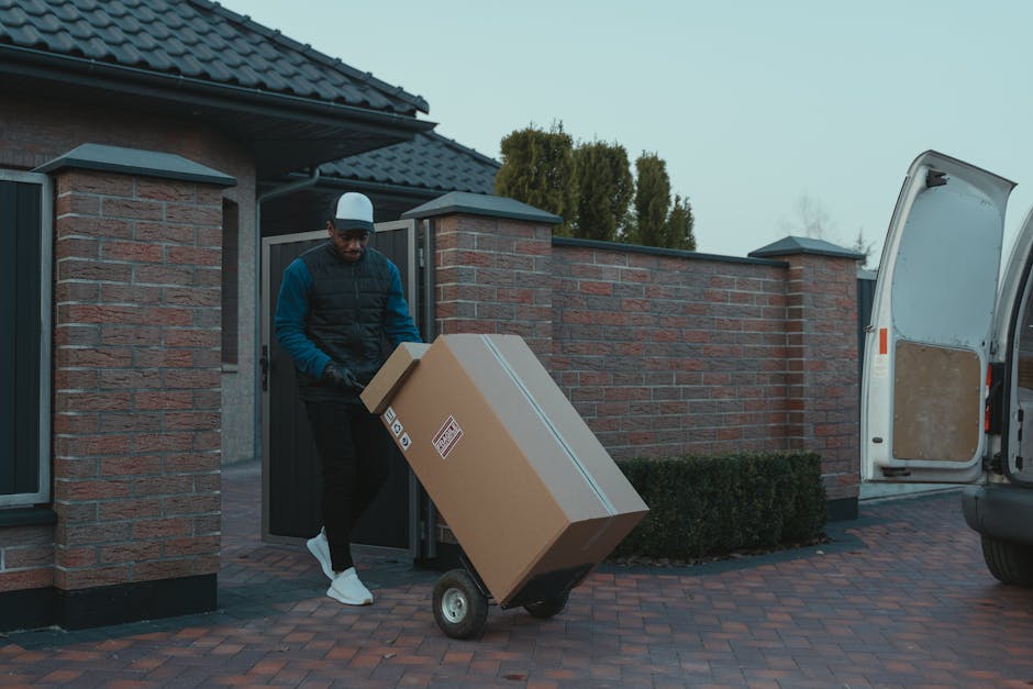 A man wearing a cap, black vest over a blue long-sleeve shirt, and dark trousers is seen loading a large cardboard box onto a black hand truck outside a residential property. The box is sealed with packing tape and has handling labels on it. The man is positioned next to a driveway paved with red bricks, with the white rear door of a van open nearby, indicating active loading or unloading. The property features a brick wall, dark roof tiles, and a closed gate, with tall trees and overcast sky visible in the background. This scene illustrates a house removal or packing and moving process facilitated by Man with Van Custom House, supporting efficient furniture transport and logistics for home relocation.
