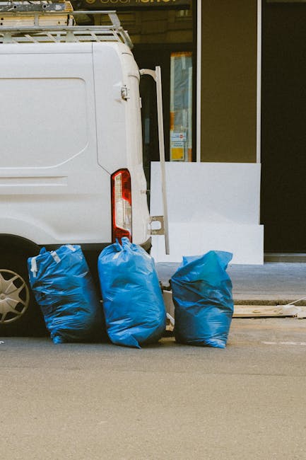 A white commercial van parked on a street outside a building with a glass storefront; at the rear of the van, three large blue rubbish bags filled with bulky waste are placed on the pavement, ready for disposal or removal. The van is positioned near an open doorway, with some white panels or furniture leaning against the building’s exterior wall nearby, indicating a home or office clearance process. The loading area shows signs of recent or ongoing loading, consistent with house removal or clearance services. The environment is well-lit, with natural daylight illuminating the scene, and the overall setting suggests a professional waste clearance or furniture removal operation, as handled by Man with Van Custom House, supporting efficient packing and moving logistics for domestic or commercial relocations.