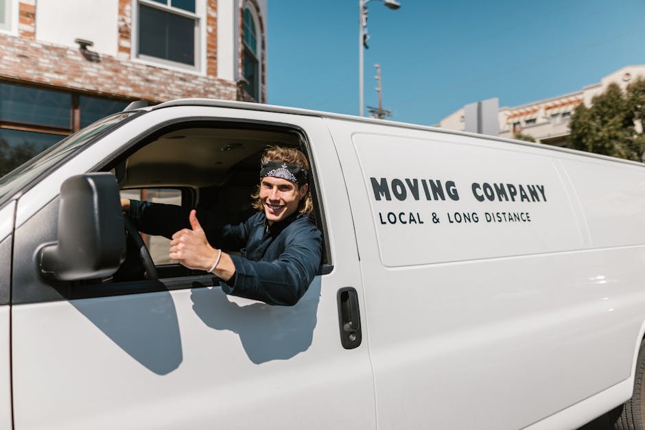 A white moving van with the company name 'MOVING COMPANY' and the words 'LOCAL & LONG DISTANCE' parked in front of a modern, two-storey residential building during daylight. Inside the vehicle, a move