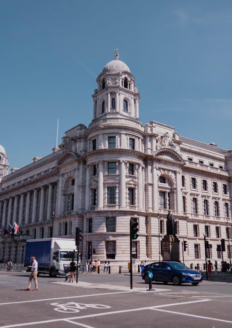 A daytime view of a large historic building with classical architectural features, including a prominent domed tower at the corner, Corinthian columns, and ornate decorative elements. The building is made of light-colored stone or concrete, with multiple windows and carved details. In the foreground, a city street scene shows a delivery van parked near the curb, pedestrians walking across the crosswalk, and a few cars driving past. Traffic lights are visible at the intersection, along with street markings for bicycles and vehicles. The scene captures a typical urban environment, potentially in the vicinity of a house removals service such as Man with Van Custom House, which might be involved in facilitating home relocation or furniture transport in the area.