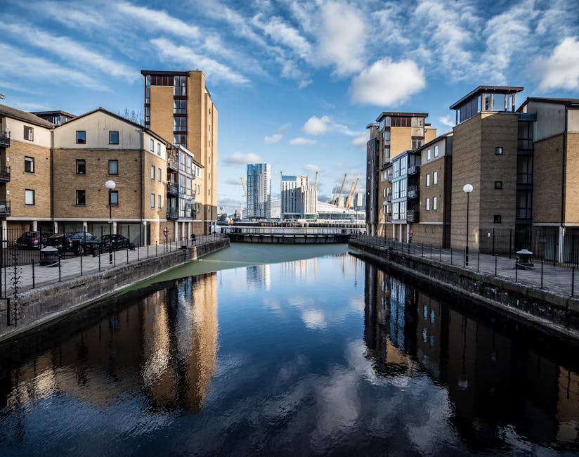 A view of a canal flanked by modern residential buildings with beige brick facades, black-framed windows, and small balconies, situated in a residential area of Custom House. Cars are parked along the canal on a paved walkway, which is lined with black metal railings and lampposts. In the background, contemporary high-rise buildings with glass exteriors and construction cranes are visible against a partly cloudy blue sky. The scene depicts an urban environment where house removals and furniture transport may occur, with the canal serving as a potential route for moving logistics and home relocation activities supported by companies like Man with Van Custom House.