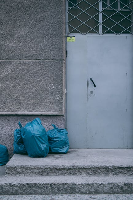 A person dressed in orange work overalls is standing inside a home with a plain white wall and grey carpeted floor. They are holding two large blue plastic garbage bags filled with waste or packing ma