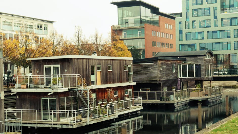 Photograph depicting two modern floating houseboats moored along a canal in the ExCeL area near Royal Docks, with contemporary high-rise buildings in the background. The houseboats feature wooden exteriors, large glass windows, and multiple outdoor decks with metal railings, some decorated with potted plants. One houseboat is positioned slightly ahead, partially visible with a staircase leading to its upper deck, while the other is parallel beside it. The scene is well-lit under natural daylight, with calm water reflecting the structures. This setting illustrates the type of residential properties involved in home relocation or furniture transport projects managed by Man with Van Custom House, demonstrating the process of packing, loading, or transporting furniture and belongings during house removals in the area.