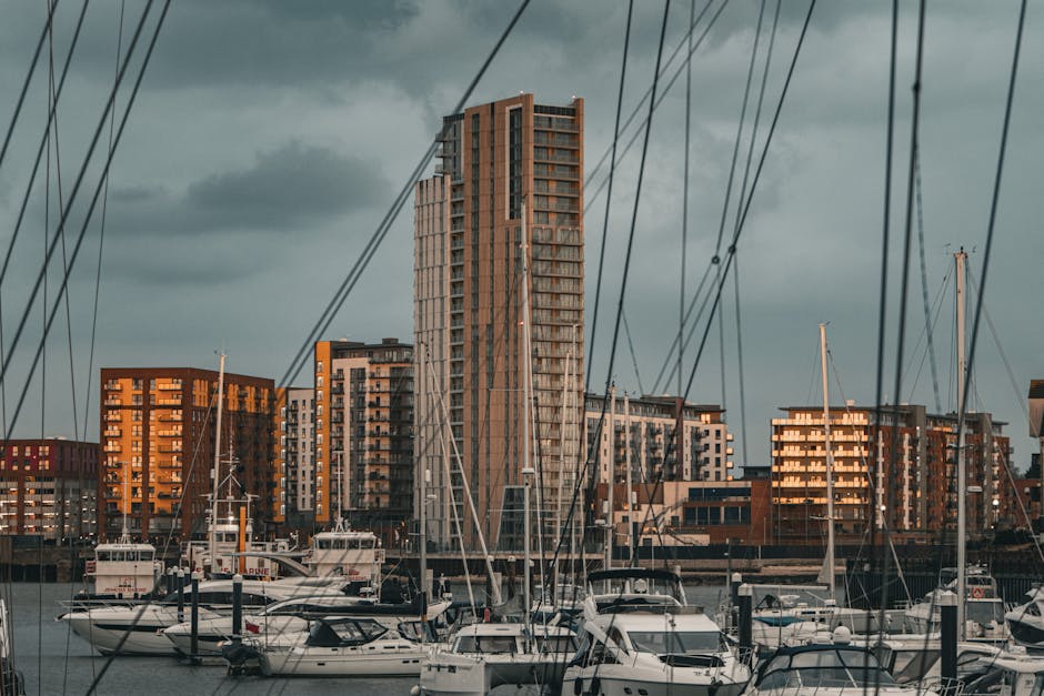 A marina scene showing several white motorboats and sailboats docked at wooden piers, with tall modern residential buildings in shades of brown, beige, and gray in the background. The boats are secured with ropes, and some have protective covers over their decks. Overhead, numerous cables and rigging lines crisscross the sky, which is overcast with dark clouds. The setting suggests a busy waterfront area, possibly in the ExCeL region near Royal Docks, with buildings reflecting the warm evening sunlight. This urban waterfront environment is relevant to house removals and furniture transport, illustrating the importance of careful planning during home relocation in areas like Custom House, where Man with Van Custom House provides professional moving services.