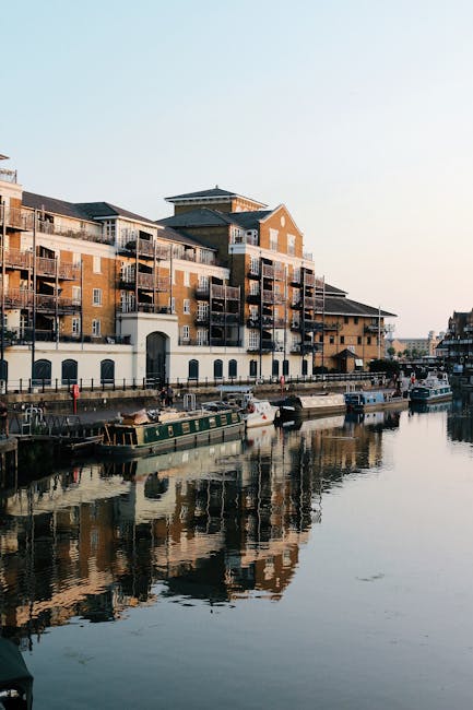 Photograph depicting two modern floating houseboats moored along a canal in the ExCeL area near Royal Docks, with contemporary high-rise buildings in the background. The houseboats feature wooden exteriors, large glass windows, and multiple outdoor decks with metal railings, some decorated with potted plants. One houseboat is positioned slightly ahead, partially visible with a staircase leading to its upper deck, while the other is parallel beside it. The scene is well-lit under natural daylight, with calm water reflecting the structures. This setting illustrates the type of residential properties involved in home relocation or furniture transport projects managed by Man with Van Custom House, demonstrating the process of packing, loading, or transporting furniture and belongings during house removals in the area.