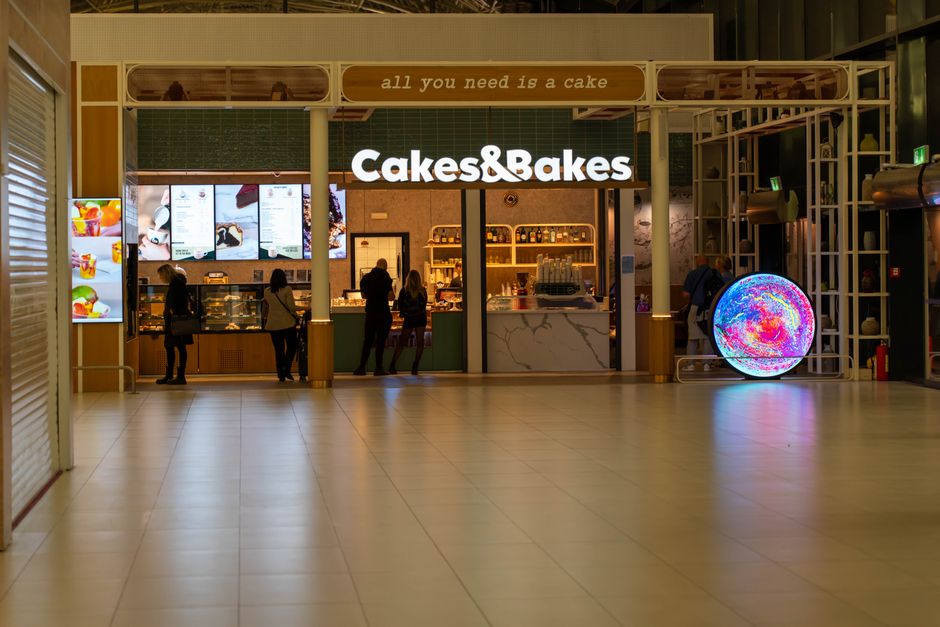 Inside a shopping mall, there is a Cakes & Bakes bakery counter with a bright illuminated sign displaying the store name. The bakery has a modern design with a marble-style counter surface and digital menu screens above it showing images and options for baked goods. Several customers are standing in front of the counter, some selecting items, while others are looking at the menus. To the right of the bakery, there is a large, colorful LED circular display on a stand, emitting vibrant, swirling lights. The floor is tiled with light-colored tiles, and the environment is well-lit with natural and artificial lighting. The first floor setting, along with the visible surroundings, suggests this image may relate to home relocation or moving services that include shopping mall or retail space logistics, which Man with Van Custom House handles as part of their removals and packing services, especially when moving belongings from commercial or retail environments.