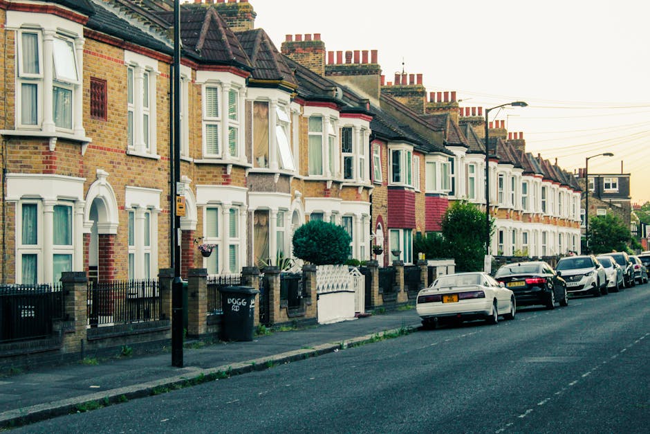 A row of multi-storey terraced houses with brick facades and bay windows, situated along a residential street in Newham, with some houses featuring white window frames and decorative elements. The pavement is lined with black metal fences, with small front gardens containing shrubs and potted plants. Several parked cars, including a white vehicle in the foreground, are present along the curb. A black wheelie bin with the label 'DOGG' is visible near one of the fences. Streetlights and power lines run above the street, and the scene is illuminated by natural light during late afternoon or early evening. This setting depicts a typical urban neighbourhood, suitable for house removals or home relocation services, highlighting residential logistics and the importance of proper packing and transport planning, as managed by Man with Van Custom House.