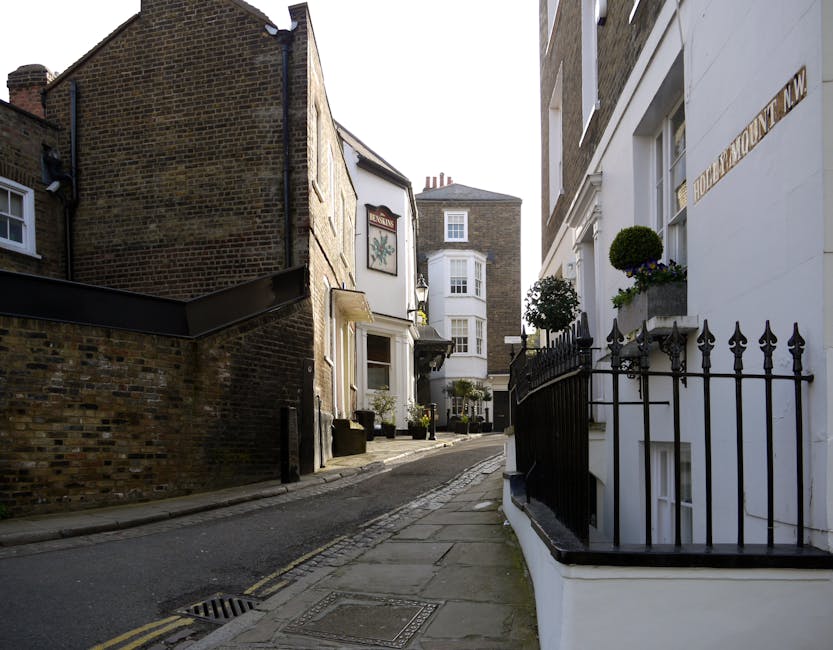 A narrow residential street during daytime featuring a mix of historic brick and white-painted houses, with potted plants and small trees on window sills and balconies, enclosed by black wrought iron railings on the right side. The pavement is uneven with concrete slabs and a drainage grate near the curb. A white building on the right, marked with a street name plaque, is in close proximity to the sidewalk. The scene captures the challenging urban environment for home relocation, with visible parts of a van or moving equipment not directly shown but implied by the context of house removals in a tight street, as handled by Man with Van Custom House. Bright natural lighting highlights the textures of brickwork and the smooth surfaces of the buildings, suggesting a clear day suitable for furniture transport and packing and moving activities.