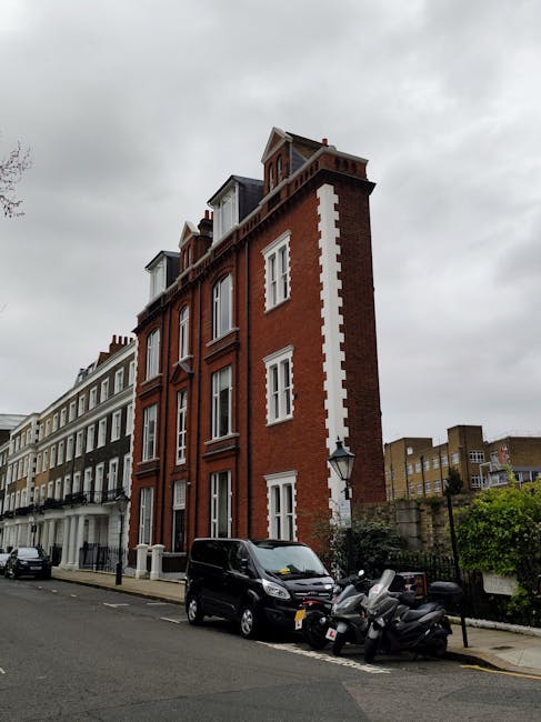 A narrow street scene outside a multi-storey red-brick residential building with white detailing, featuring large windows and a small rooftop structure. In the foreground, a black van is parked on the pavement alongside several parked scooters, all near the building's entrance. The street is lined with similar terraced houses, and the sky overhead is cloudy, indicating overcast weather. This setting likely depicts a home relocation process occurring in an urban area, with the presence of parking and moving equipment suggesting recent or upcoming furniture transport or packing and moving activities, as would be handled by a professional removals company such as Man with Van Custom House.
