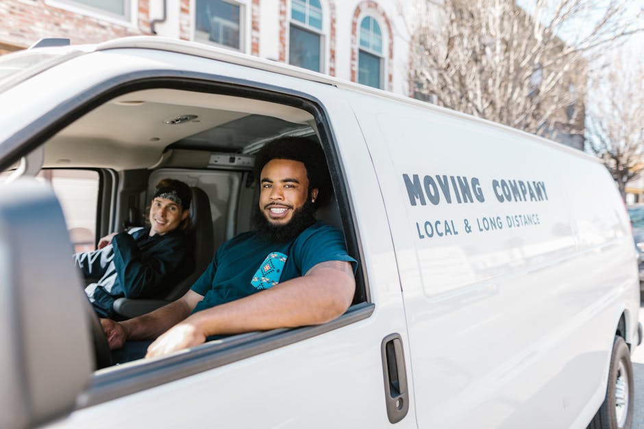 A white moving van parked outdoors on a paved street during daytime, featuring the words 'Moving Company' and 'Local & Long Distance' printed on the side. Inside the van's open side door, two men are visible: one sitting in the driver’s seat, smiling, with short curly hair and a blue T-shirt, and another in the passenger seat, also smiling, wearing a black jacket and a cap. Several cardboard boxes wrapped in plastic and packing materials are arranged inside the van, indicating an active home relocation or furniture transport process. The scene captures the loading or unloading phase with the vehicle positioned near residential buildings, under natural daylight, illustrating a professional and organized moving service provided by [COMPANY_NAME].