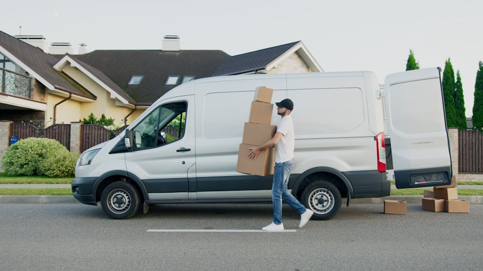 A man wearing a white t-shirt, jeans, and a black face mask loads three medium-sized cardboard boxes into the back of a white panel van parked on a residential street. The van's rear doors are open, revealing a spacious cargo area. A few additional boxes are placed on the ground beside the van. The scene is set in front of modern houses with dark roofs, light-colored walls, and tall green bushes, under a bright sky with daylight illuminating the area. The man is carrying the boxes from the pavement towards the van, indicating a home relocation or packing process. The surroundings include a neatly maintained garden and a low wooden fence, suggesting a quiet suburban environment. [COMPANY_NAME], which provides removals and moving services, appears to be involved in this furniture transport activity as part of a house move, emphasizing efficient loading and transport logistics.